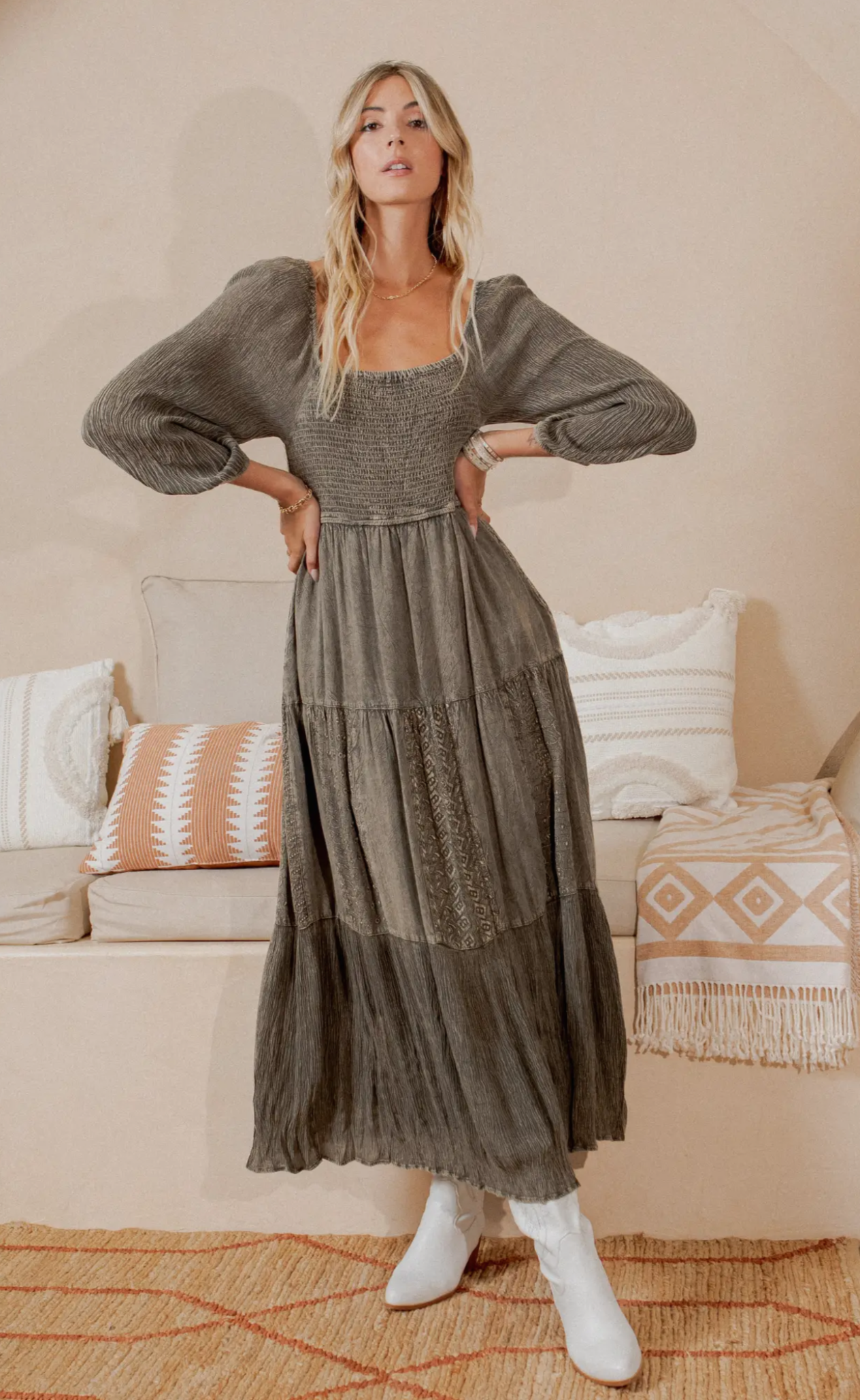 A woman standing in an indoor setting wearing a mid-length, embroidered, square neck dress in a neutral color.